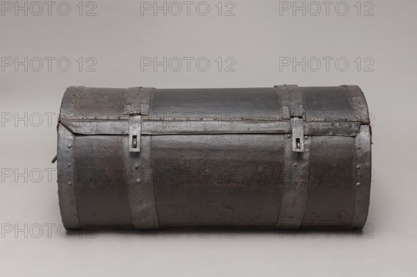 Trunk, A TUBE in the shape of a cylinder truncated at the base, covered with thick leather and upholstered with four steel straps, fastened with steel heads, with U-shaped handles on the sides of the body. The lid is closed with two hasps, attached to strap hinges. The connection between the body and the lid is protected from water by a leather apron. The side straps on the outside have semicircular cutouts, folded. The interior of the trunk is lined with gray canvas. The lid, when opened, is held on the sides with leather strips. 17th/18th century, Poland, piece of furniture, height 46 cm, width 104.5 cm, depth 51.5 cm., A cylindrical chest with a domed lid made of dark metal with visible rivets and metal bands, showing a worn and slightly textured surface in shades of dark brown and black with areas of dull gray; a few vertical metal straps wrap around the body and lid, each secured with rivets and featuring rectangular metal fastenings, and the piece rests on a smooth light gray bac...