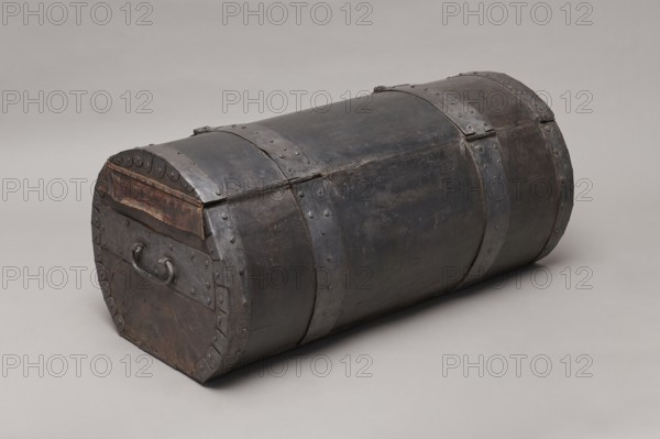 Trunk, deposit, pre-war collection — A TUBE in the shape of a cylinder truncated at the base, covered with thick leather and upholstered with four steel straps fastened with steel heads, with U-shaped handles on the sides. The lid is closed with two hasps attached to strap hinges. The connection between the body and the lid is protected from water by a leather apron. The side straps on the outside have semicircular cutouts, folded. The interior is lined with gray canvas. The lid, when opened, is held on the sides with leather strips. 17th/18th century, Poland, furniture; H 46 cm, W 104.5 cm, D 51.5 cm., A cylindrical chest lying on its side with a domed lid, made of dark metal with visible rivets and metal bands; the surface shows worn and slightly rusty patches and textured imperfections. The chest has a flat end with a metal handle attached and decorative rounded rivet plates along the edge. The colors visible are dark gray, black, brown, and subtle rust tones, set against a neutral ...