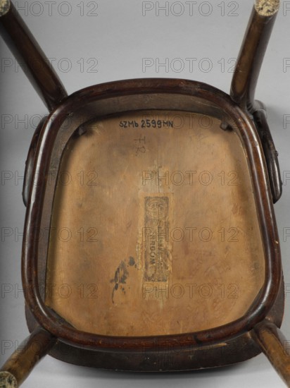 Chair, Johann Kohn & Cie Radom; bentwood furniture factory; late 19th century?-1939. Bentwood chair with an openwork backrest and a trapezoidal seat made of extruded plywood with rounded corners. Profiled, straight front legs, slightly diagonally inclined at the bottom; rear legs arch outward, merging into the sideways-flared backrest frames. The legs are joined at the top with a hoop repeating the outline of the seat. U-shaped connectors between the backrest frames and the seat frame. The backrest has two horizontal slats bent in the back line, the upper one wider, with three slats between them. Circa 1900–1914, Radom. Seating furniture; height 89.5 cm, width 39.5 cm, depth 49 cm., A wooden chair seen from underneath with a rounded rectangular underside and curved wooden frame, showing a worn light brown wooden panel inside a darker brown wooden rim and legs; a few areas of darker brown scuff marks and worn spots appear on the panel and frame, and there are traces of adhesive residue ...