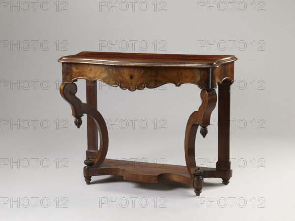 Console, Biedermeier and Louis Philippe styles, mahogany-veneered console on four angular legs mounted on a platform with a wavy outline. Front legs strongly curved; rear legs straight, flattened, with bases and capitals. Entire piece polished. Platform supported by four low turned legs. Lower sections of the legs composed of channel sections decorated at top and bottom with knobbed pinnacles. Wide frame repeating concave-convex shape of the top, rounded at front corners, wavy-cut at bottom, framed with a half-cylinder; front frame forms the face of the drawer. Top has a profiled edge connected to the frame with cube side panels. Drawer has two rows of compartments inside; small front-corner compartments with rosewood covers. Drawer secured by a single-bolt mortise lock. Dated 1835–1850. Piece of furniture table. Height 82 cm, width 107.2 cm, depth 45 cm., A wooden table with a polished brown finish sits against a pale gray background; the tabletop has a curved rectangular shape with a...