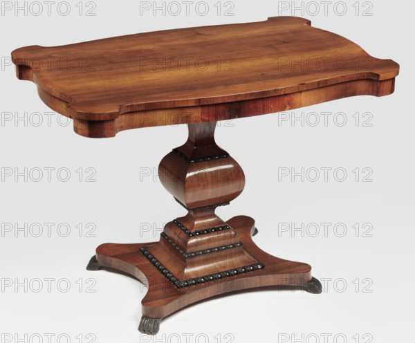 Table, Biedermeier style, animal paws, rosewood. Table with a rectangular top with a wavy cut, rounded corners, supported by a quadrilateral profiled shaft mounted on a square plinth with concave sides and four legs in the form of animal paws. The shaft is strongly narrowed below and above the knop, with the lower and upper parts widening towards the platform and the cube connecting it to the top; framed with strips carved with an astragal motif and blackened like the secondary legs. The frame repeats the outline of the top, with a drawer on one of the long sides. The top is narrowed at the bottom by two cross strips acting as side guides for the drawer; attached to the frame with nine sideboards. The whole piece is varnished. Date: 1840–1850. Place of origin: Vilnius . Furniture: table. Dimensions: height 78.7 cm; width 108.8 cm; depth 73 cm., A wooden table with a curved rectangular top and a carved pedestal base, showing a polished grain in shades of brown and warm reddish-brown, wi...