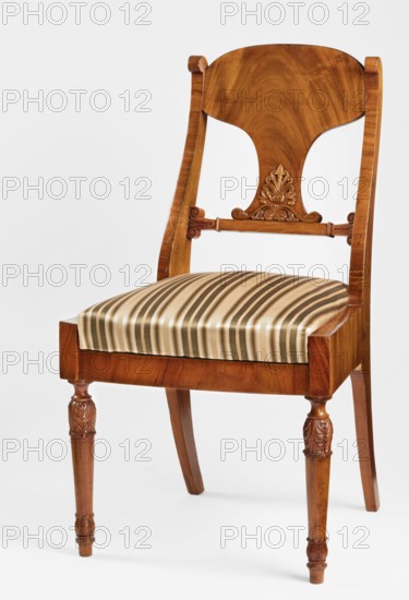Chair, acanthus ornament, Biedermeier style, palmettes ornament. Mahogany chair with a padded seat removable cushion and a board backrest. Straight front legs, turned in the form of a baluster; sabre-shaped rear legs. Trapezoidal seat, slightly bulging at the front. Wide side frames embrace the seat cushion. The cushion rests on a pine frame, upholstered in green-striped fabric. The backrest consists of angular, S-shaped side stiles, a lower crossbar, and a rounded central board placed between them, narrowed at the bottom, straight in line with the back. The backrest stiles are widened in the lower section, ending at the top with a forward-curved volute. Polished frame enriched with carving — acanthus leaves on the upper part of the front legs, antemion and symmetrical S-shaped beams at the bottom of the backrest boards, scrollwork at the ends of the backrest crossbars. Date: 1830–1840, Warsaw, Poland. Seating furniture; furniture. Dimensions: height 92 cm, width 51.6 cm, depth 56.5 cm...