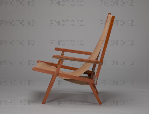 Deckchair, Polish Timber Industry, Czerski and Jakimowicz Lviv; furniture factory; 1929–1939,  furniture factory author; lounge, beach, and garden folding deckchair constructed from two different sized rectangular frames: backrest smaller and seat larger, connected by hinges, with canvas stretched over them. Frame elements except the turned rear leg connector and the doubled front seat stile are angular. Seat frame supported on its shorter sides by two folding legs connected by a connector, the front extended into armrest supports. Armrests have hinges adjustable in three positions. Salmon-colored canvas with vertical orange stripes framed by narrow blue stripes; canvas attached to undersides of the upper backrest frame and the rear seat frame, sandwiched between the two front seat frames. Originally equipped with a separate footrest. 1938–1939, Lviv; piece of seating furniture. Height 84.2 cm; width 58.5 cm; depth 115.5 cm., A wooden folding chair with a canvas seat and back is shown ...