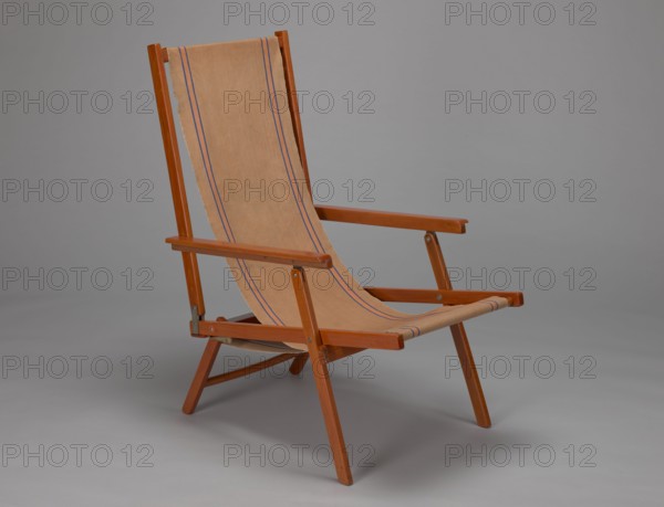 Deckchair, Polish Timber Industry, Czerski and Jakimowicz furniture factory; 1929–1939. Folding deckchair constructed from two different sized rectangular frames: the backrest smaller and the seat larger, connected by hinges, with canvas stretched over them. Frame elements except the turned rear leg connector and the doubled front seat stile are angular. The seat frame is supported on its shorter sides by two folding legs connected by a connector, the front extended into armrest supports. The armrests have hinges adjustable in three positions. Salmon-colored canvas fabric with vertical orange stripes framed by narrow blue stripes; attached to the underside of the upper backrest frame and the rear seat frame, sandwiched between the two front seat frames. Originally equipped with a separate footrest. 1938–1939, Lviv; seating furniture deckchair. Height 84.2 cm; width 58.5 cm; depth 115.5 cm., A wooden folding chair with a tan fabric sling seat and back is shown against a plain gray backg...