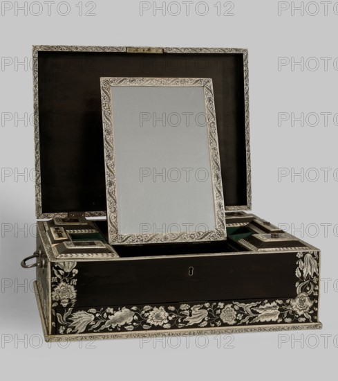 Travel dressing table, Baroque style, ca. 1750, Vizagapatam, India. Ebony cuboid with a lifting lid and a front bottom drawer, richly inlaid with flowers and plant twigs engraved in bone and set with black paint. Inside: a tilting mirror, five compartments on each side six with lids. Mirror frame and lids inlaid with bone with plant and floral twigs. Three compartments in the drawer. Steel swan neck handles on the sides. Travel/hygiene furniture. Height 17 cm, width 41 cm, depth 30.5 cm., A decorative box with its lid open showing an inner rectangular panel propped upright, all surfaces predominantly dark brown with ornate light-colored floral and foliate inlays and borders; the propped panel has an intricate light-colored frame and a pale central surface, the interior lid is dark, and the box interior includes recessed compartments with green fabric linings and light-colored trim; metal hardware in a muted metallic tone appears at the side and front keyhole area, and the box sits agai...
