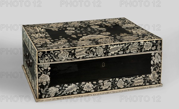 Travel dressing table, ebony cuboid with lifting lid and a front bottom drawer, richly inlaid with flowers and plant twigs in engraved bone set with black paint. Inside: a tilting mirror, five compartments on each side six with lids; mirror frame and lids inlaid with bone with plant and floral twigs. Three compartments in the drawer. Steel swan neck handles on the sides. Decorated with flowers and plant ornamentation in a Baroque style. Ca. 1750, Vizagapatam, India. Travel/hygiene furniture. Dimensions: H 17 cm, W 41 cm, D 30.5 cm., A rectangular wooden box with a hinged lid decorated with intricate floral and foliate patterns in contrasting light and dark tones; the box surface is predominantly black with ornate cream-colored floral designs covering the top, front, sides, and edges, forming borders and scattered blossoms and leaves. The front shows a recessed panel bordered by floral bands, and there are a few keyhole-shaped openings visible on the front face. The left side has a dark...