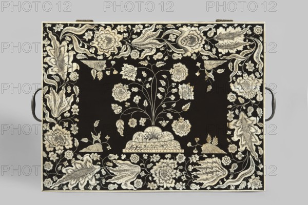 Travel dressing table, flowers, plant ornament, Baroque style, Lubomirski family, flower bushes ornament. Travel dressing table in ebony, cuboid, with a lifting lid and a drawer at the bottom of the front, richly inlaid with flowers and plant twigs in engraved bone, set with black paint. Inside: a tilting mirror, five compartments on each side six with lids. The mirror frame and lids are inlaid with bone with plant and floral twigs. Three compartments in the drawer. Steel handles on the sides swan neck. Ca. 1750, Vizagapatam, India. Travel/hygiene furniture. Height 17 cm, width 41 cm, depth 30.5 cm., A rectangular object with a black surface decorated with an intricate cream-colored floral pattern fills the image; a central stylized plant with stems and blossoms rises from a scalloped base, surrounded by a border of large ornate flowers, leaves, and curling tendrils, with a pair of dark metal handles visible at the left and right edges.