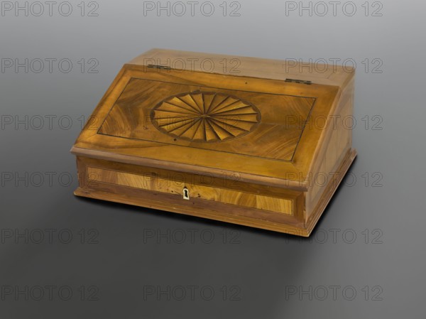 Traveling secretary, classicism style, platter ornament. TRAVELING SECRETARY with straight walls, with a diagonally inclined flat lid, ash veneered, inlaid. On all walls and the lid are geometric fields adapted to the outline, enclosed by a frame, filled with patterned ash and edged with black and light veining. On the lid is an oval reserve with a platter motif with tan arms. Inside, a central recess enclosed by an arcade, two drawers on each side in two rows. Bone lock key; steel, mortise-and-applied, single-bolt, latch lock. Brass hinges, greyish. 18th/19th century, Poland. Furniture, writing furniture. Height 17.1 cm, width 40.5 cm, depth 30.6 cm., A rectangular wooden box with a sloped lid sits on a smooth gray surface; the box is a warm golden-brown with visible wood grain and a darker brown trim, and the sloped top features a carved circular fan-like rosette at its center surrounded by inset panels with grain patterns; the front shows a small keyhole plate in a lighter metal and...