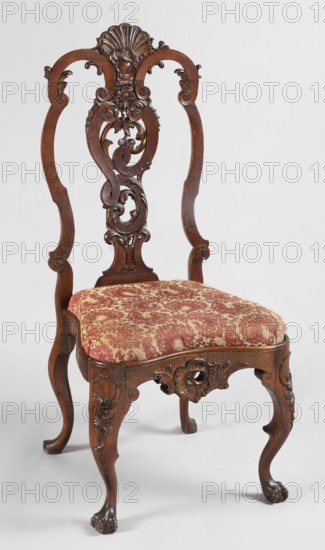 Chair, Carved chair with a padded seat inserted cushion and a high backrest filled by an openwork central splat, supported by four cabriole legs — front legs more strongly curved and widened at the top, terminating in ball-and-claw feet; rear legs with slipper feet. Back frame slightly rounded; front frame concave in the center and wavy-cut at the bottom, like the knees of the legs, decorated with rocaille motifs. Central splat asymmetrical, with the outline of a vase formed by ribbon, embellished with acanthus leaves and flowers. Upper backrest frame consists of two channel-shaped sections and a carved shell, with small channel-shaped acanthus leaves on the sides. Ornamentation: animal paws, acanthus, shells, plant decoration, volutes. Dated 1745–1760, Netherlands. Seating furniture; height 109.3 cm, width 50 cm, depth 49.5 cm., A wooden chair with an ornate carved back featuring swirling and foliate motifs and a shell-like crest at the top, a padded seat upholstered in patterned fabr...