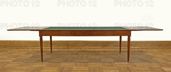 Table, Biedermeier style, attributed to around 1830–1837, rectangular table with drop-down flaps on the short sides; supported by four turned, tapering legs with knuckles at the top. Main top plate lined in the middle with grey cloth, framed by a mahogany frame connected to the frame by sideboards. Flaps veneered with mahogany. Wooden sliders with slatted flap supports glued to the underside of the main top part on the short sides. Profiled legs with a roll-up at the top and pairs of rings below and halfway up; frame stained mahogany, polished. Maker associated with Józef Pilsudski 1867–1935. Origin: Warsaw. Function: furniture, table. Dimensions: height 78.4 cm; width 221 cm; depth 96 cm., A wooden table with an extended top and a green inset surface, standing on turned wooden legs on a parquet wooden floor against a light-colored wall with a wooden baseboard; visible colors include brown, green, beige, and white.