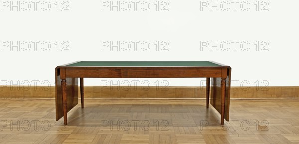 Table, Biedermeier style, attributed to, rectangular table with drop-down flaps on the short sides; supported by four turned, tapering legs with knuckles at the top. Main top plate lined in the middle with grey cloth, framed by a mahogany frame; connected to the frame by sideboards. Flaps veneered with mahogany. Wooden sliders with slatted flap supports glued to the underside of the main top part on the short sides. Profiled legs with a roll-up at the top and pairs of rings below and halfway up; frame stained mahogany, polished. Inscription/association: Pilsudski, Józef 1867–1935. Dated 1830–1837, Warsaw. Furniture; table. Dimensions: height 78.4 cm, width 221 cm, depth 96 cm., A wooden table with a green top sits centered against a pale off-white wall and a light brown wooden baseboard, standing on a patterned light brown parquet floor; the table has a warm medium-brown finish with turned legs and drop leaves visible at the sides, and the visible colors include green, medium brown, li...