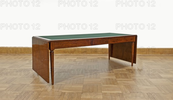 Table, Biedermeier style, associated with Józef Pilsudski 1867–1935. Rectangular table with drop-down flaps on the short sides; supported by four turned, tapering legs with knuckles at the top. Main top plate lined in the middle with grey cloth, framed by a mahogany frame and connected to the frame by sideboards. Flaps veneered with mahogany. Wooden sliders with slatted flap supports glued to the underside of the main top part on the short sides. Profiled legs with a roll-up at the top and pairs of rings below and halfway up; frame stained mahogany, polished. Dated 1830–1837, origin Warsaw. Furniture, table; height 78.4 cm, width 221 cm, depth 96 cm., A wooden table with a green top sits on a light brown parquet floor against a pale off-white wall with a beige baseboard; the table has a polished medium brown finish with visible wood grain, turned legs at the front, flat side panels at each end, and a thin pale trim around the edge of the green surface.