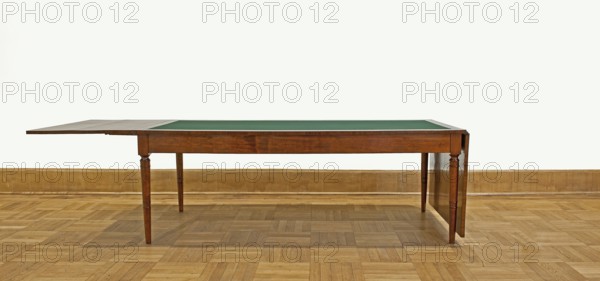 Table, Biedermeier style, attributed to, associated with Józef Pilsudski 1867–1935. Rectangular table with drop-down flaps on the short sides; supported by four turned, tapering legs with knuckles at the top. Main top plate lined in the middle with grey cloth, framed by a mahogany frame; connected to the frame by sideboards. Flaps veneered with mahogany. Wooden sliders with slatted flap supports glued to the underside of the main top part, on the short sides. Profiled legs with a roll-up at the top and pairs of rings below and halfway up; frame stained mahogany, polished. Dated 1830–1837, Warsaw. Furniture; table. Dimensions: height 78.4 cm, width 221 cm, depth 96 cm., A wooden table with a green inset surface stands on a parquet wood floor against a plain light-colored wall with a wooden baseboard; the table has a medium brown finish, turned legs, and an extended leaf on one side, and the scene shows colors including brown, green, tan, and off-white.