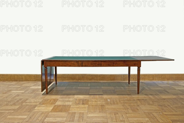 Table, Biedermeier style 1830–1837, rectangular table with drop-down flaps on the short sides; supported by four turned, tapering legs with knuckles at the top. Main top plate lined in the middle with grey cloth, framed by a mahogany frame connected to the frame by sideboards. Flaps veneered with mahogany. Wooden sliders with slatted flap supports glued to the underside of the main top part on the short sides. Profiled legs with a roll-up at the top and pairs of rings below and halfway up; frame stained mahogany, polished., Origin: Warsaw. Function: furniture, table. Dimensions: height 78.4 cm, width 221 cm, depth 96 cm., A long wooden table with a green top surface and a wooden extension on one end stands on a patterned wooden floor against a plain light-colored wall with a wooden baseboard; the table has turned wooden legs and a visible wood grain in rich brown tones, while the floor shows varying shades of light to medium brown and the wall appears off-white.