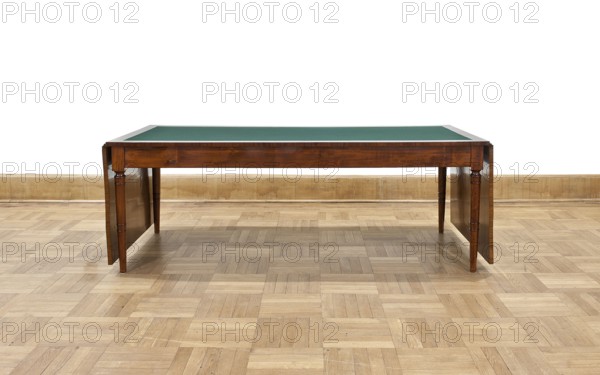 Table, Biedermeier style; rectangular table with drop-down flaps on the short sides; supported by four turned, tapering legs with knuckles at the top. Main top plate lined in the middle with grey cloth, framed by a mahogany frame; connected to the frame by sideboards. Flaps veneered with mahogany. Wooden sliders with slatted flap supports glued to the underside of the main top part on the short sides. Profiled legs with a roll-up at the top and pairs of rings midway; frame stained mahogany, polished. Inscription/association: Pilsudski, Józef 1867–1935. Date: 1830–1837. Place of origin: Warsaw. Function: furniture/table. Dimensions: height 78.4 cm; width 221 cm; depth 96 cm. Creator: unknown., A wooden table with a green top sits on a patterned wooden floor against a pale wooden baseboard and white wall; the table has a rich brown finish with turned legs and drop leaves on either side, and the visible colors include brown, green, tan, beige, and white.