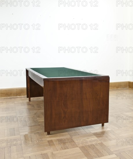Table, Biedermeier style, associated with Józef Pilsudski 1867–1935. Rectangular table with drop-down flaps on the short sides; supported by four turned, tapering legs with knuckles at the top. The main top plate is lined in the middle with grey cloth, framed by a mahogany frame; connected to the frame by sideboards. The flaps are veneered with mahogany. Wooden sliders with slatted flap supports are glued to the underside of the main top part, on the short sides. Profiled legs with a roll-up at the top, below and halfway up with pairs of rings and a frame stained mahogany, polished. Dated 1830–1837, Warsaw. Furniture, table; height 78.4 cm, width 221 cm, depth 96 cm., A wooden table with a dark brown finish and a green top sits on a light wood parquet floor against a white wall with a light brown baseboard; the table shows wood grain on its sides and has small round legs, the room appears brightly lit and mostly empty, with visible colors including brown, green, white, and light tan.