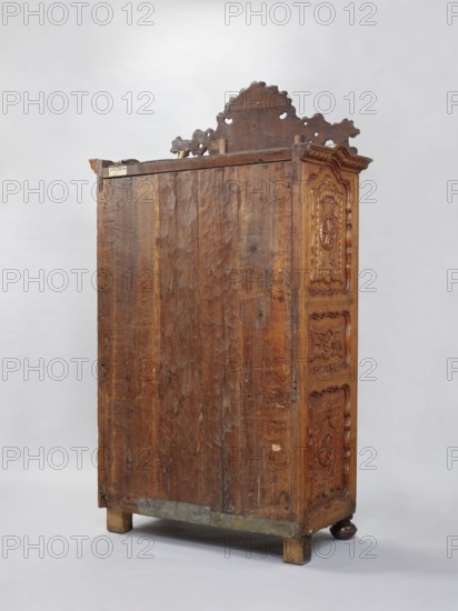 Wardrobe Armoire, late 18th century, France. Rococo style with rocaille ornament, plant decoration, and rosettes. Cuboid wardrobe closed with double doors, richly carved and topped with a decorative board featuring an asymmetrical Rococo cartouche framed by rocaille motifs. Supported by four low legs — front legs slightly flattened spheres, rear legs block-shaped. Sides of the body and door leaves framed with three flat-carved panels bearing stylized plant motifs and rosettes on a star-punched background; frames symmetrically cut inside with a wavy profile. Board backs set in a rebate, fastened with dowels and nails; saw marks present. Interior: three narrowed central shelves with wooden galleries the fourth shelf is the upper surface of a compartment containing two drawers at the bottom; the upper shelf rests on openwork consoles with rocaille motifs. Shelves secured to the sides with large-diameter pins ~15 mm. Steel fittings; eyelet hinges secondary; push-pull lock not preserved. Fu...