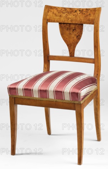 Chair, Biedermeier style, Biedermeier chair with a trapezoidal, padded seat and a rectangular openwork backrest curved outward in an S-shape. Prismatic legs, arched outward; the rear legs merge into the backrest stiles. Straight frame. Upholstered seat on springs. A wide board tops the backrest; a strip at the bottom, with a cup-shaped middle board between them. Date: 1825–1835. Place of origin: Warsaw. Type: seating furniture, furniture. Dimensions: height 86 cm, width 46 cm, depth 45.5 cm., A wooden chair with a warm honey-brown finish and visible wood grain and knots, featuring a curved backrest with a decorative central splat, a rectangular seat upholstered in striped fabric with alternating deep red and cream bands, a narrow decorative trim along the seat edge in a gold tone, gently tapered legs in the same honey-brown wood, and a plain light gray background behind the chair.