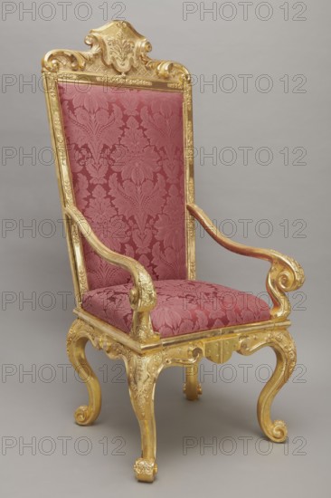 Travel armchair, late Baroque type, carved and gilded, with a high flat backrest, cabriole legs that arc into a wavy-cut bottom frame, and S-shaped armrests whose supports end in a common volute. Trapezoidal seat and backrest cushions, reupholstered in red damask imitating large-scale Baroque fabrics. Recessed, removable backrest upholstery after removing the upper frame mounted on steel pins and decorated with acanthus leaves, a checkered pattern, and a cartouche filled with an acanthus finial. Frame decorated with palmettes and rosettes impressed in chalk-glue; legs framed with a half-cylinder. Hinges connecting front and rear frames made of steel, primed and gilded on the outside. Dated 1710–1725; probable origin Germany. Seating piece, height 175 cm, width 73 cm, depth 64 cm., A tall ornate chair with a carved gilded wooden frame and a richly patterned upholstery in shades of deep red and rose; the high back and seat display a damask-like floral design, and the armrests curve into ...