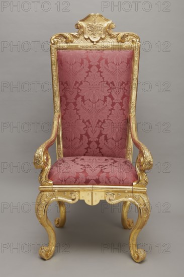 Travel armchair, late Baroque type, carved and gilded, with a high flat backrest, on cabriole legs that arc into a wavy-cut lower frame, and with S-shaped armrests whose supports end in a common volute. Trapezoidal seat and backrest cushions reupholstered in red damask imitating large-scale Baroque fabrics. Backrest upholstery is recessed and removable after removing the upper frame, which is mounted on steel pins and decorated with acanthus leaves, a checkered pattern, and a cartouche filled with an acanthus finial. Frame decorated with palmettes and rosettes impressed in chalk-glue; legs framed with a half-cylinder. Hinges connecting front and rear frames are steel, primed and gilded on the outside. Travel armchair; carved, gilded wood and textiles; ca. 1710–1725; Germany attributed. Height 175 cm; width 73 cm; depth 64 cm., An ornate high-backed chair with a carved gilded wooden frame and a patterned upholstery in a deep red color; the frame shows gold color with intricate scrollwor...