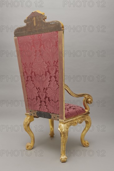 Travel armchair of the late Baroque type, carved and gilded, with a high flat backrest on cabriole legs that arc into the wavy-cut frame at the bottom, S-shaped armrests and supports ending in a common volute. Trapezoidal seat and backrest cushions, reupholstered in red damask imitating large-scale Baroque fabrics. The backrest upholstery is recessed and removable after removing the upper frame, which is mounted on steel pins and decorated with acanthus leaves, a checkered pattern, and a cartouche filled with an acanthus finial. Frame decorated with palmettes and rosettes impressed in chalk-glue; legs framed with a half-cylinder. Hinges connecting the front and rear frames are steel, primed and gilded on the outside. Late Baroque, 1710–1725; probable origin Germany. Seating furniture travel armchair. Dimensions: height 175 cm, width 73 cm, depth 64 cm., A tall ornate chair seen from the back with a high rectangular backrest upholstered in patterned deep red fabric framed by wooden trim...