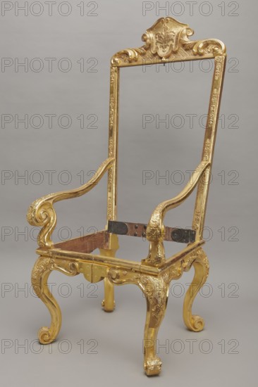 Travel armchair, late Baroque type, carved and gilded, with a high flat backrest on cabriole legs that arc into a wavy-cut bottom frame; S-shaped armrests with supports ending in a common volute. Trapezoidal seat and backrest cushions reupholstered in red damask imitating large-scale Baroque fabrics. Recessed, removable backrest upholstery after removing the upper frame mounted on steel pins and decorated with acanthus leaves, a checkered pattern, and a cartouche filled with an acanthus finial. Frame decorated with palmettes and rosettes impressed in chalk-glue; legs framed with a half-cylinder. Hinges connecting front and rear frames of steel, primed and gilded on the outside. Travel, volutes, floral decoration; Baroque style. Made circa 1710–1725, Germany . Seating piece, height 175 cm, width 73 cm, depth 64 cm., A tall ornate chair frame with a gilded finish, showing a decorative crest at the top and carved scrollwork along the uprights and arms, with curved armrests that end in spi...