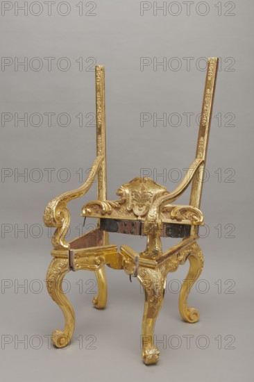 Travel armchair, late Baroque type, carved and gilded, with a high flat backrest, cabriole legs that arc into a wavy-cut bottom frame, S-shaped armrests with supports ending in a common volute. Trapezoidal seat and backrest cushions, reupholstered in red damask imitating large-scale Baroque fabrics. The backrest upholstery is recessed and removable after removing the upper frame, which is mounted on steel pins and decorated with acanthus leaves, a checkered pattern, and a cartouche filled with an acanthus finial. Frame decorated with palmettes and rosettes impressed in chalk-glue; legs framed with a half-cylinder. Hinges connecting front and rear frames are steel, primed and gilded on the outside. Travel function; carved wood, gilding, upholstery. Dated 1710–1725. Origin: Germany, Height 175 cm; width 73 cm; depth 64 cm., A gilded ornate chair frame with a high back structure stands against a neutral gray background; the frame has curving armrests and cabriole legs with carved scroll f...