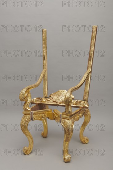Travel armchair, travel, volutes, floral decoration, Baroque, travel armchair of the late Baroque type, carved, gilded, with a high, flat backrest, on cabriole legs that arc into the wavy-cut frame at the bottom, and with S-shaped armrests and their supports ending in a common volute. Trapezoidal seat and backrest cushions, reupholstered in red damask imitating large-scale Baroque fabrics. The backrest upholstery is recessed and removable after removing the upper frame, which is mounted on steel pins and decorated with acanthus leaves, a checkered pattern, and a cartouche filled with an acanthus finial. The frame is decorated with palmettes and rosettes impressed in chalk-glue, and the legs are framed with a half-cylinder. The hinges connecting the front and rear frames are made of steel, primed and gilded on the outside. 1710–1725, Germany, piece of furniture, seating piece; height 175 cm, width 73 cm, depth 64 cm., A partially assembled ornate chair frame with a gilded finish stands ...