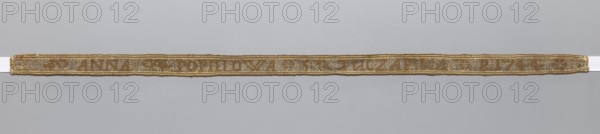 Gallon, galloons haberdashery; woven inscription: brown letters on a silver background; parallel to the edge, two white-brown stripes on each side; 1744; Poland; fabric; haberdashery; width 1.4 cm; length 41 cm., A narrow woven band lies horizontally across a pale gray background, showing a textured surface with a metallic sheen; the band has a warm golden color with areas of muted brown and pale beige, and it is edged with a slightly darker golden border.
