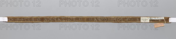 Gallon, haberdashery; woven inscription in brown letters on a silver background; parallel to the edge, two white-brown stripes on each side; 1744; Poland; fabric; width 1.4 cm; length 41 cm., A narrow woven band displayed horizontally against a light gray background, showing a long strip of textured fabric in brown and tan tones with subtle variations in shade and a slightly darker woven pattern along its length; the band has a thin tan border on its top and bottom edges and a few small attached labels at one end that include small patches of white and a small piece of light orange, with a short loop of pale string nearby.