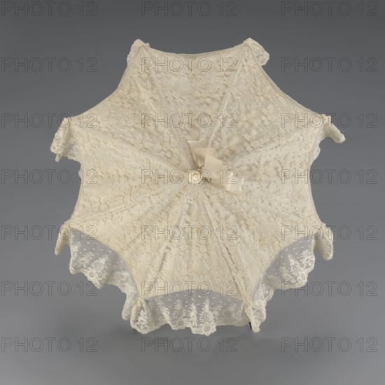 Umbrella, women's umbrella made of white lace. Canopy trimmed with cream silk at the bottom, concealing the metal structure and providing a backdrop for the lace when unfolded. Bone handle carved with floral motifs and folds in half. c. 1850–1875, France. Length 66 cm; diameter 57 cm. Categories/subjects: umbrellas, fashion, lace, women's fashion, 19th-century fashion accessories., A delicate lace parasol is shown from above, with an octagonal shape and scalloped edges, made of fine off-white and cream lace with floral embroidery and dotted mesh; a small bow and fabric rosette in similar cream tones sit near the center, and several lace ruffles and decorative tabs extend from the edges, all photographed against a smooth neutral gray background.