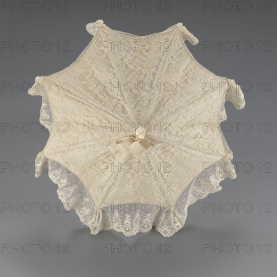 Women's umbrella made of white lace. Its canopy is trimmed with cream silk at the bottom, which conceals the metal structure and, when unfolded, provides a backdrop for the lace covering. The bone handle is carved with floral motifs and folds in half. c. 1850–1875, France. Length 66 cm; diameter 57 cm., A delicate lace parasol in cream tones is shown from above against a gray background, with a scalloped edge of lace and floral lace patterns radiating across its canopy, a gathered fabric rosette and ribbon at the center, and small curved fabric-covered tips at the outer points.