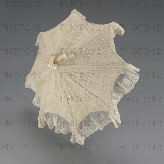 Women's umbrella made of white lace. Its canopy is trimmed with cream silk at the bottom, which conceals the metal structure and, when unfolded, provides a backdrop for the lace covering. The bone handle is carved with floral motifs and folds in half. c. 1850–1875, France. Length 66 cm; diameter 57 cm. Categories: umbrellas; fashion; lace; women's fashion; 19th century fashion accessories; fashion accessories; lace varietals., An open lace parasol with a scalloped edge displayed against a plain gray background, the parasol fabric appearing off-white or cream with a delicate floral lace pattern and dotted mesh, a small bow and ornamental cluster at the center where the ribs meet, and the lace edges showing intricate floral motifs and slight translucency.