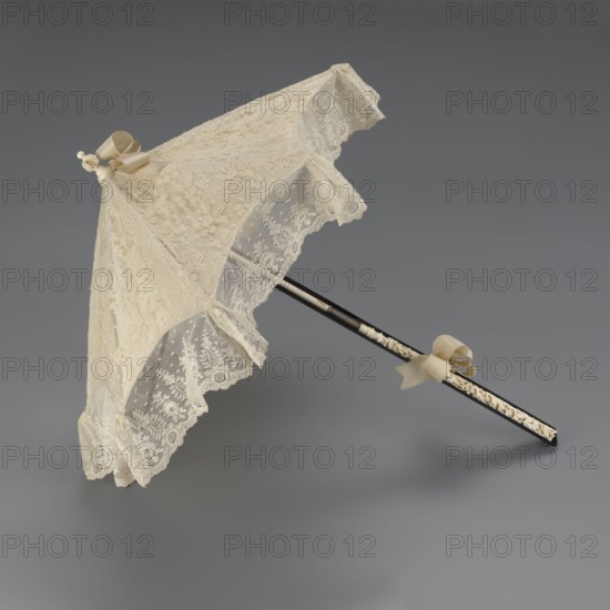 Women's umbrella made of white lace. Its canopy is trimmed with cream silk at the bottom, which conceals the metal structure and, when unfolded, provides a backdrop for the lace covering. The bone handle is carved with floral motifs and folds in half. c. 1850–1875, France. Length 66 cm; diameter 57 cm. Materials/attributes: umbrellas, fashion, lace, women's fashion, 19th century fashion accessories., A delicate lace parasol in shades of cream and ivory rests against a neutral gray background, featuring a scalloped lace canopy with floral and dotted patterns, a few decorative bows in a pale beige attached near the top and along the handle, and a slender dark shaft with a carved or inlaid lighter decorative band running along part of its length; the lace edges form a ruffled trim and the overall appearance is ornate and finely detailed.