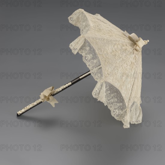 Women's umbrella made of white lace. Its canopy is trimmed with cream silk at the bottom, which conceals the metal structure and, when unfolded, provides a backdrop for the lace covering. The bone handle is carved with floral motifs and folds in half. c. 1850–1875, France. Length 66 cm, diameter 57 cm. Fashion accessory; 19th-century women's fashion., A lace parasol with an open canopy rests on a smooth gray background; the canopy is an off-white or cream color with delicate floral lace and scalloped edges, and it has a matching cream bow near the top and a few cream ribbons tied along the shaft; the shaft is dark, appearing black or very dark brown, with an inner section that shows a lighter, carved or textured cream-colored detail; overall colors visible include cream, off-white, black or dark brown, and gray.