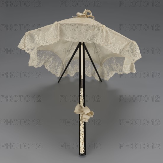 Women's umbrella made of white lace. Its canopy is trimmed with cream silk at the bottom, which conceals the metal structure and, when unfolded, provides a backdrop for the lace covering. The bone handle is carved with floral motifs and folds in half. c. 1850–1875, France. Length 66 cm; diameter 57 cm. Keywords: umbrellas, fashion, lace, women's fashion, 19th century fashion accessories, fashion accessories., A lace parasol in shades of cream and off-white with a scalloped lace edge and floral lace patterns, supported by a black pole and black ribs, with a decorative cream bow tied near the top and another cream bow near the handle, shown against a smooth gray background.