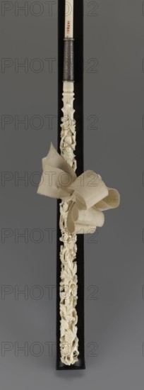 Women's umbrella made of white lace. Its canopy is trimmed with cream silk at the bottom, which conceals the metal structure and, when unfolded, provides a backdrop for the lace covering. The bone handle is carved with floral motifs and folds in half. c. 1850–1875, France. Length 66 cm, diameter 57 cm., A long slender object displayed vertically against a dark background with a carved pale ivory section featuring an intricate openwork floral and foliate design, a smooth pale handle area, and a textured metallic band near the top; a soft fabric bow in beige tones is tied around the carved section, and the overall color palette includes shades of pale ivory, beige, dark gray, and black.