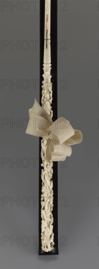 Women's umbrella made of white lace. Its canopy is trimmed with cream silk at the bottom, which conceals the metal structure and, when unfolded, provides a backdrop for the lace covering. The bone handle is carved with floral motifs and folds in half. c. 1850–1875, France. Length 66 cm; diameter 57 cm. Keywords: umbrellas; fashion; lace; women's fashion; 19th century fashion accessories., A narrow vertical carved object in off-white with intricate floral and foliate openwork along its length, set against a black rectangular backing and a gray background; a fabric bow in a beige tone is tied around the object near its upper third, with layered loops and visible woven texture; the carved surface shows smooth areas and detailed raised petals and leaves, and the overall palette includes off-white, beige, black, and gray.