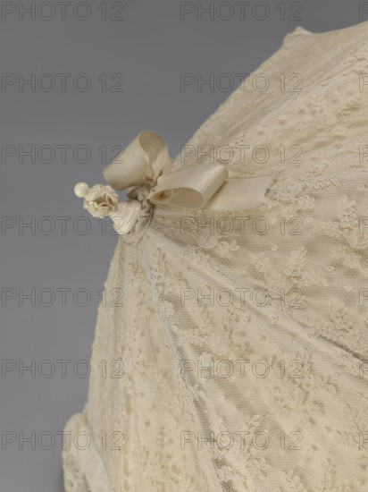 Women's umbrella made of white lace. Its canopy is trimmed with cream silk at the bottom, which conceals the metal structure and, when unfolded, provides a backdrop for the lace covering. The bone handle is carved with floral motifs and folds in half. c. 1850–1875, France. Length 66 cm; diameter 57 cm., A close-up view of a cream-colored lace fabric gathered and draped, featuring a satin ribbon bow in a similar cream tone tied near the top, with a small decorative finial attached that appears ivory and rounded; the lace shows delicate floral patterns and a fine netting texture, and the background is a smooth mid-gray.