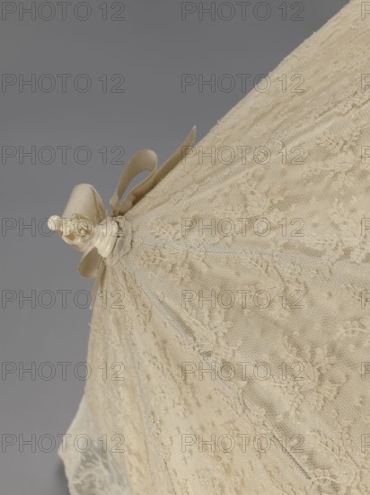 Women's umbrella made of white lace. Its canopy is trimmed with cream silk at the bottom, which conceals the metal structure and, when unfolded, provides a backdrop for the lace covering. The bone handle is carved with floral motifs and folds in half. c. 1850–1875, France. Length 66 cm; diameter 57 cm., The image shows a close-up of a pale cream lace umbrella or parasol against a gray background, featuring delicate floral lace patterns and a sheer netting overlay, with a decorative ivory finial and a matching cream ribbon bow near the top; visible colors include cream, ivory, and gray.