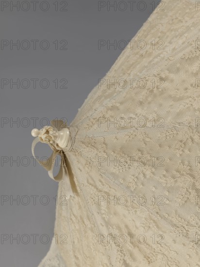 Women's umbrella made of white lace. Its canopy is trimmed with cream silk at the bottom, which conceals the metal structure and, when unfolded, provides a backdrop for the lace covering. The bone handle is carved with floral motifs and folds in half. c. 1850–1875, France. Length 66 cm, diameter 57 cm., A close-up of a lace fabric showing a delicate floral pattern in an off-white or cream color with a small cream-colored decorative finial and ribbon tied to the fabric; the background is a smooth gray, and the lace displays a repeating textured motif of tiny floral and leaf details.