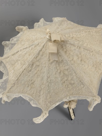 Women's umbrella made of white lace. Its canopy is trimmed with cream silk at the bottom, which conceals the metal structure and, when unfolded, provides a backdrop for the lace covering. The bone handle is carved with floral motifs and folds in half. c. 1850–1875, France. Length 66 cm; diameter 57 cm., An ornate lace parasol shown from above with a scalloped edge and intricate floral lace overlay in cream and ivory tones, a central decorative finial topped with a small ribbon in matching cream, delicate sheer mesh panels embroidered with floral motifs and tiny dotted textures, a tied bow near the handle in cream, and a slim dark handle partially visible beneath the canopy against a neutral gray background.