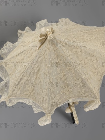 Umbrella. Women's umbrella made of white lace with a canopy trimmed with cream silk at the bottom that conceals the metal structure and, when unfolded, provides a backdrop for the lace covering. The bone handle is carved with floral motifs and folds in half. c. 1850–1875, France. Length 66 cm; diameter 57 cm., umbrellas; fashion; lace; women's fashion; 19th century fashion accessories; fashion accessories; varietals; lace., An ornate lace parasol seen from above, predominantly cream and beige in color with a delicate floral lace pattern across its canopy, scalloped lace edges, a decorative bow near the top with small fabric flowers, and a slender handle partially visible beneath the canopy in darker tones.