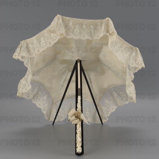 Umbrella, women's umbrella made of white lace. Canopy trimmed with cream silk at the bottom, concealing the metal structure and, when unfolded, providing a backdrop for the lace covering. Bone handle carved with floral motifs and folds in half. c. 1850–1875, France. Length 66 cm; diameter 57 cm., A delicate parasol with a scalloped lace canopy in shades of cream and off-white is open and viewed from beneath, showing a translucent fabric interior with a soft beige tone and a decorative lace border; the ribs and shaft are black with a contrasting ivory-toned section near the top, and a carved ivory-colored handle with an attached cream ribbon bow rests at the base against a neutral gray background.