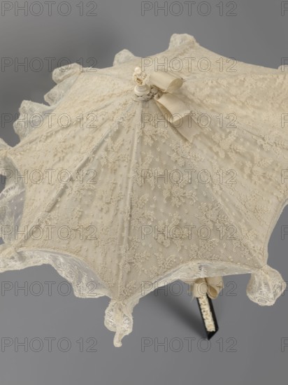 Women's umbrella made of white lace. Its canopy is trimmed with cream silk at the bottom, which conceals the metal structure and, when unfolded, provides a backdrop for the lace covering. The bone handle is carved with floral motifs and folds in half. Circa 1850–1875, France. Length 66 cm; diameter 57 cm., An open lace parasol with delicate floral embroidery and a scalloped lace edging in shades of cream and off-white sits against a neutral gray background, showing a central finial topped with a small decorative element and a tied ribbon, and a handle partially visible with similar cream lace detailing.