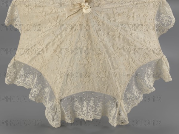 Umbrella. Women's umbrella made of white lace; canopy trimmed with cream silk at the bottom, concealing the metal structure and, when unfolded, providing a backdrop for the lace covering. Bone handle carved with floral motifs and folds in half. c. 1850–1875, France. Length 66 cm; diameter 57 cm., umbrellas, fashion, lace, women's fashion, 19th century fashion accessories., A delicate lace parasol canopy displayed against a neutral background, primarily in cream and off-white tones, featuring an overall dotted mesh with embroidered floral motifs, scalloped edges with denser lace panels, a small fabric bow and a round decorative element near the top, and visible folds and gentle draping creating soft shadows in pale beige and gray.