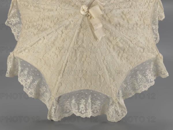 Women's umbrella made of white lace. Its canopy is trimmed with cream silk at the bottom, which conceals the metal structure and, when unfolded, provides a backdrop for the lace covering. The bone handle is carved with floral motifs and folds in half. c. 1850–1875, France. Length 66 cm; diameter 57 cm., A close-up view of a lace parasol showing delicate floral lace panels and scalloped lace edging, with a ribbon and small rosette near the top; colors visible include cream and beige on the lace and ribbon, with a gray background.