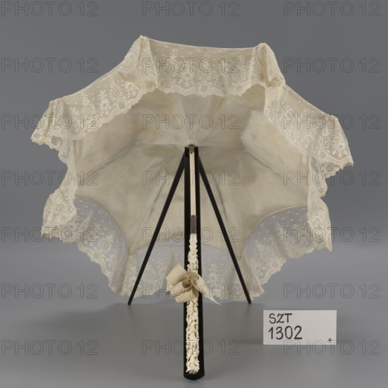 Women's umbrella made of white lace. Its canopy is trimmed with cream silk at the bottom, which conceals the metal structure and, when unfolded, provides a backdrop for the lace covering. The bone handle is carved with floral motifs and folds in half. c. 1850–1875, France. Length 66 cm; diameter 57 cm. Materials: lace, silk, bone, metal. Classification: fashion accessory; umbrella; women's fashion, 19th century., A lace-trimmed parasol displayed open with a light beige canopy and decorative cream lace edging, supported by black ribs and a central black shaft with a carved cream decorative section and a cream ribbon bow attached; the background is gray and the surface beneath is gray.
