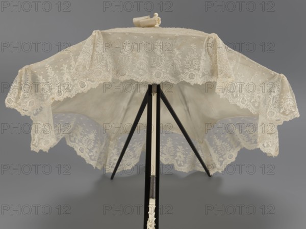 Women's umbrella made of white lace. Its canopy is trimmed with cream silk at the bottom, which conceals the metal structure and, when unfolded, provides a backdrop for the lace covering. The bone handle is carved with floral motifs and folds in half. c. 1850–1875, France. Length 66 cm; diameter 57 cm., An opened parasol displayed from beneath with a scalloped lace canopy in cream and off-white tones, featuring floral and geometric lace patterns and a sheer fabric underlayer; a central slender shaft in black and cream extends downward with a carved cream handle at the end; supporting ribs in black radiate from the shaft; the background is a smooth medium gray.