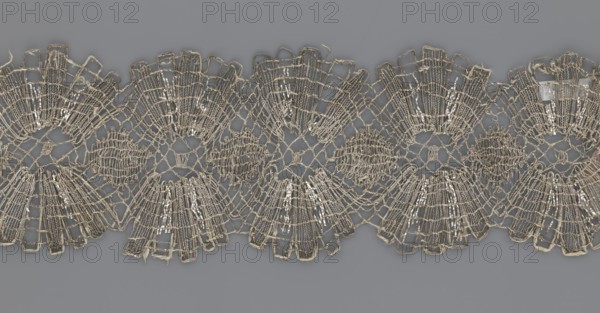 Lace fragment, lace, provenance unknown, metal threads, 18th century, Europe, lace fabric, width 8.2 cm, length 27.5 cm., A close-up of a decorative lace trim displayed against a flat background, showing a repeating scalloped pattern with open net-like sections and fan-shaped motifs; the lace appears metallic with a pale gold or silver sheen and some off-white threads, while the background is a muted gray.